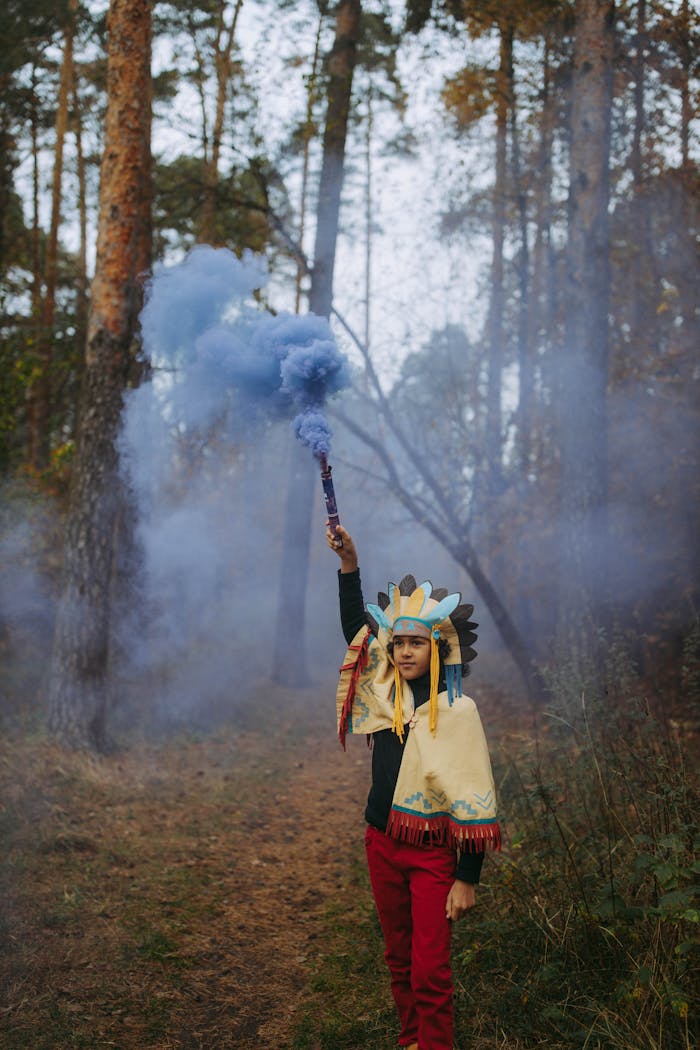 pexels-photo-5604962 A young teen in cultural attire holds a blue smoke bomb amidst tall trees in a serene forest setting.